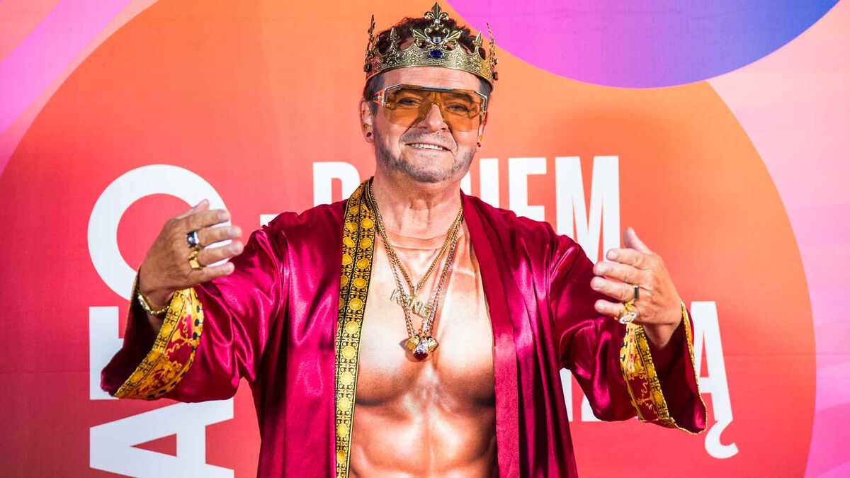 Piotr Pręgowski pozuje przed kolorową ścianką, ubrany w bordowy satynowy płaszcz, koronę, okulary z pomarańczowymi szkłami oraz liczną biżuterię. Aktor ma wyciągnięte przed siebie ręce, a na koszulce widoczny jest nadruk przedstawiający umięśniony tors. W tle znajduje się wielobarwna ścianka z dużymi białymi napisami i okrągłymi elementami graficznymi.