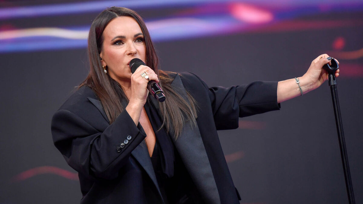 Kasia Kowalska występuje na scenie z mikrofonem w dłoni i stoi obok statywu mikrofonowego. Artystka ma na sobie czarną marynarkę, a kadr obejmuje górną część sylwetki podczas koncertu. W tle widoczne jest sceniczne oświetlenie w odcieniach fioletu i różu.