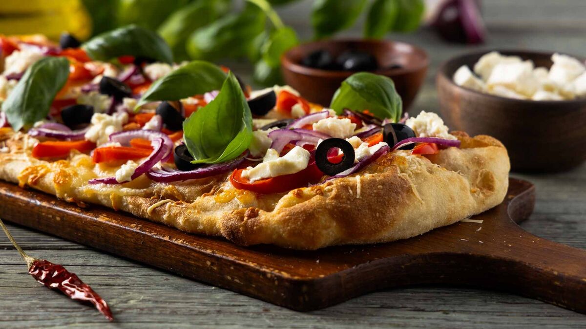 Na drewnianej desce leży pizza z dodatkiem czerwonej cebuli, czarnych oliwek, kawałków sera i świeżej bazylii. W tle widoczne są miseczki z oliwkami i serem oraz składniki użyte do przygotowania dania.