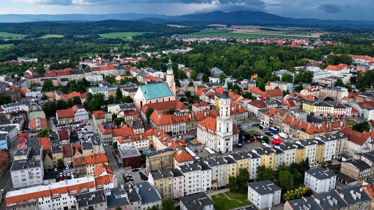 Widok z lotu ptaka przedstawia centrum miasta z rynkiem, ratuszem i zabytkowymi kamienicami. W centralnej części kadru znajduje się kościół z zielonym dachem, a wokół rozciąga się zwarta zabudowa oraz tereny zielone. W oddali widoczne są lasy, pola i pasmo gór pod zachmurzonym niebem.