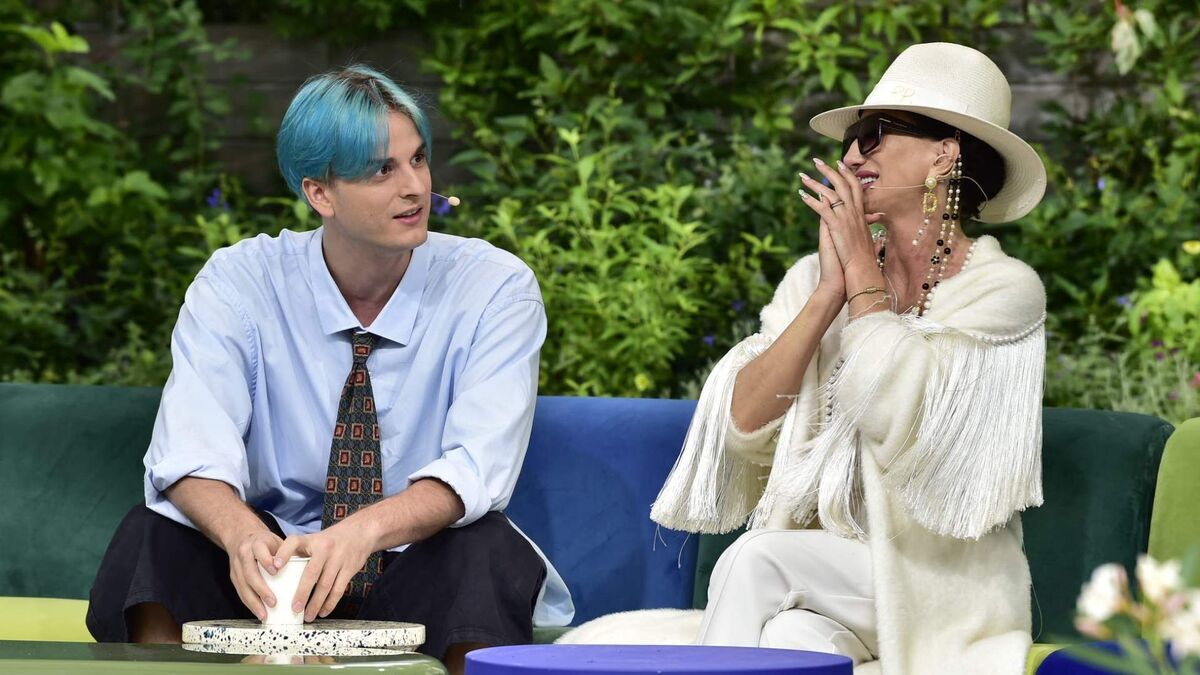 Tribbs i Justyna Steczkowska siedzą obok siebie na kanapie podczas programu telewizyjnego. Tribbs ma niebieskie włosy, jasnoniebieską koszulę i wzorzysty krawat, a Justyna Steczkowska ubrana jest na biało, ma kapelusz, ciemne okulary i długie ozdobne kolczyki. W tle widoczna jest dekoracja z zielonych roślin.