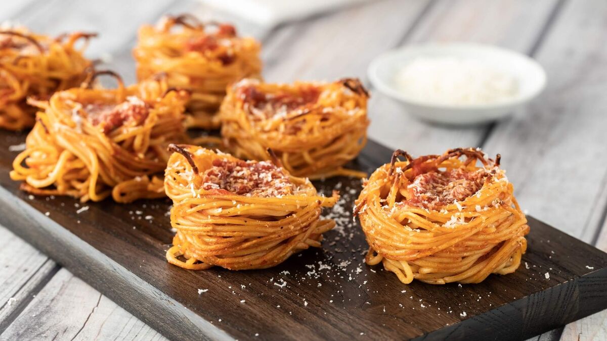 Na drewnianej desce ułożono kilka gniazd z makaronu spaghetti zapieczonych na złoto. W środku każdego „koszyczka” widać sos pomidorowy z mięsnym farszem, a całość jest oprószona startym serem. W tle stoi mała miseczka z dodatkowym serem na jasnym, drewnianym blacie.