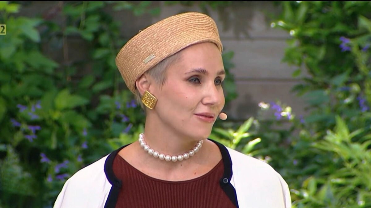 Maria Krotewicz podczas występu w studiu telewizyjnym lub programu na żywo. Ma na sobie jasny kapelusz, perłowy naszyjnik, duże złote kolczyki oraz mikrofon nagłowny. W tle widoczne są zielone rośliny stanowiące element scenografii.
