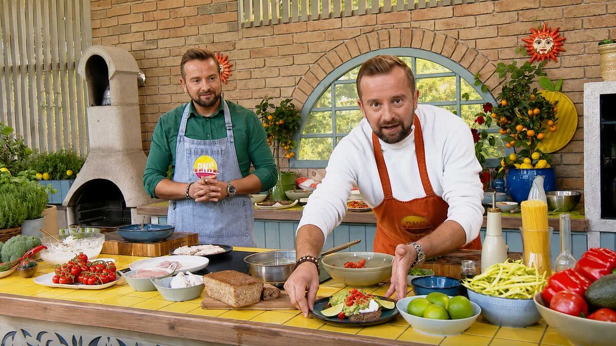 Dwóch mężczyzn stoi przy kuchennym blacie zastawionym produktami i naczyniami, a jeden z nich podaje talerz z przygotowaną potrawą. Obaj mają na sobie fartuchy, jeden pomarańczowy, a drugi jasnoniebieski. W tle znajdują się ceglana ściana, łukowe okno, piec oraz donice z roślinami.