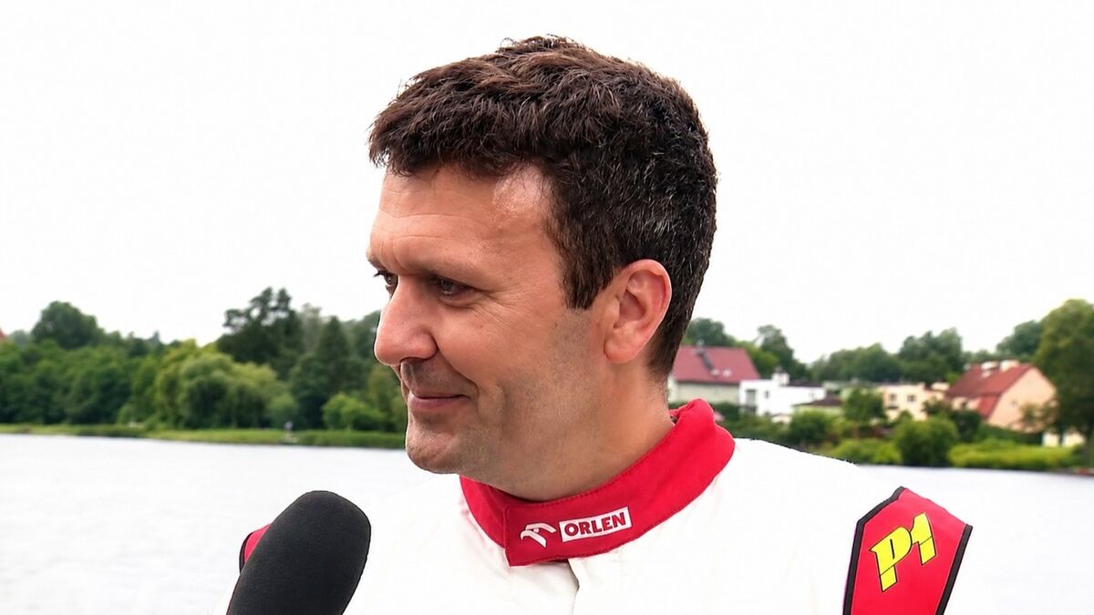 Bartłomiej Marszałek stoi bokiem do kamery i udziela wywiadu do mikrofonu nad brzegiem jeziora. Ma na sobie biało-czerwony kombinezon z logo ORLEN i oznaczeniem „P1” na ramieniu. W tle widoczne są drzewa, zabudowania oraz spokojna tafla wody.