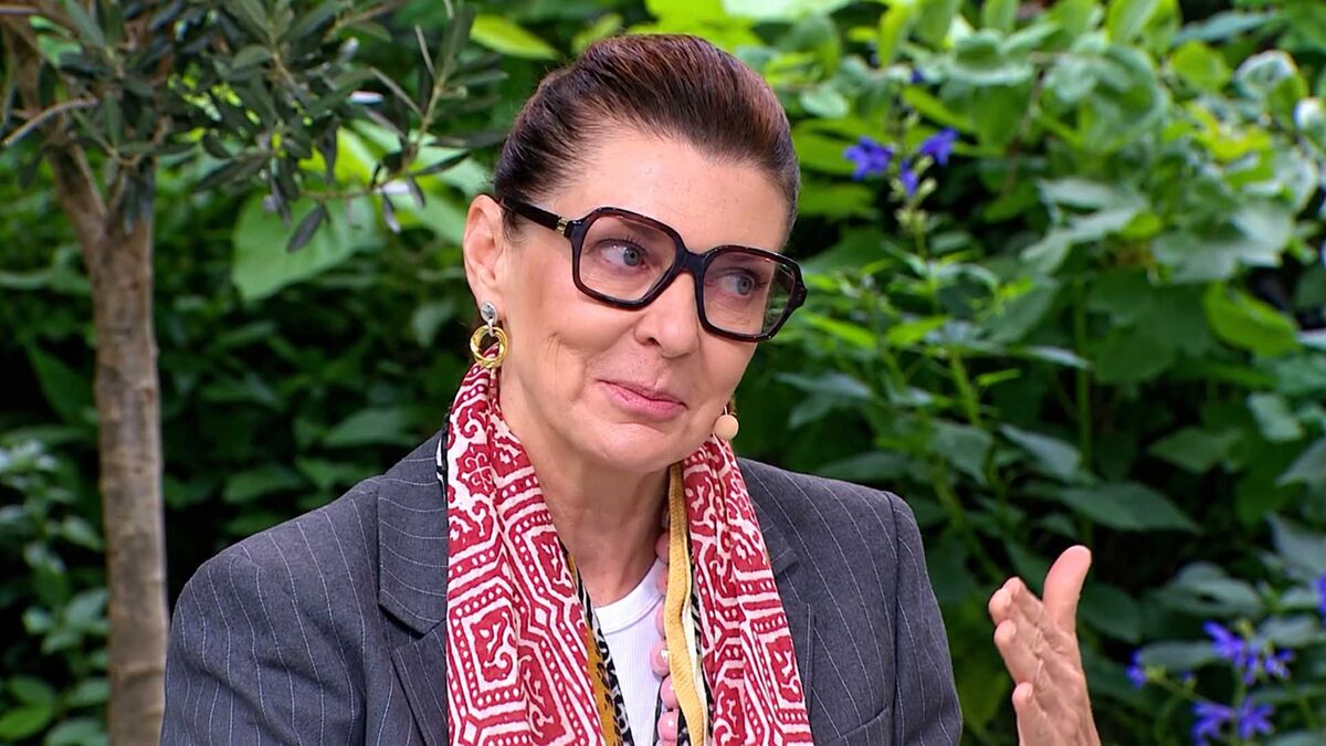 Katarzyna Kalicińska w dużych ciemnych okularach i szarej marynarce w prążki gestykuluje uniesioną dłonią. Ma włosy zaczesane do tyłu, złote kolczyki oraz czerwono-biały wzorzysty szal. W tle widoczne są gęste zielone rośliny, drzewo i drobne fioletowe kwiaty.
