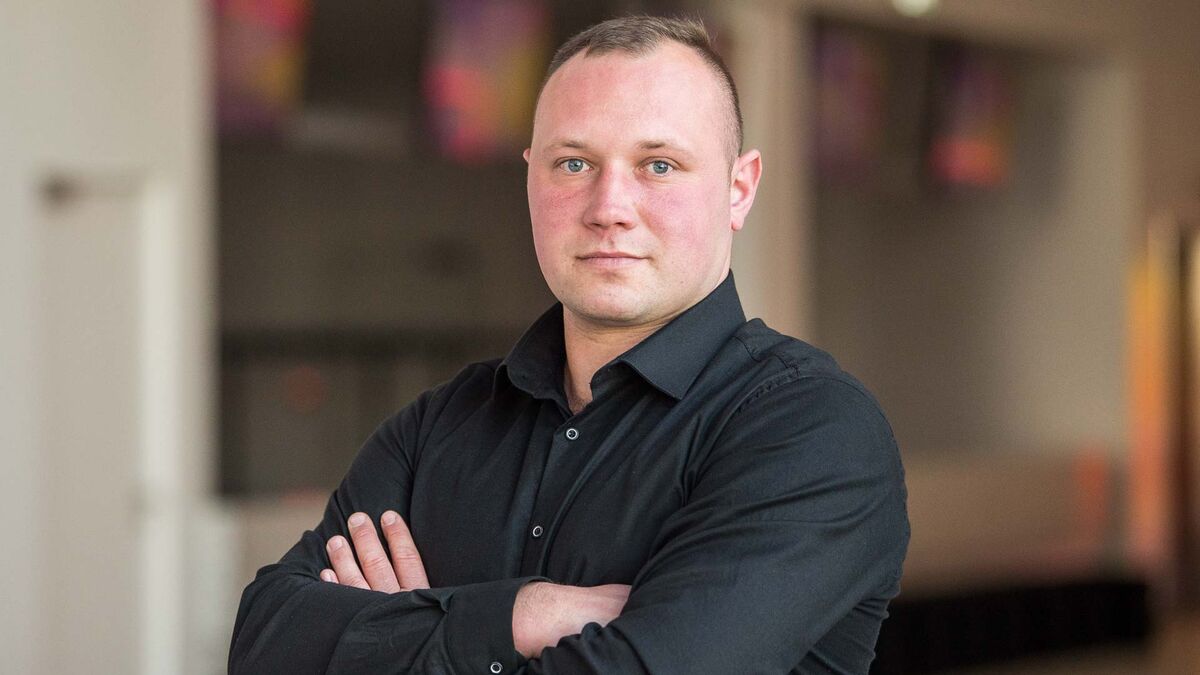 Mariusz z programu „Rolnik szuka żony” stoi ze skrzyżowanymi rękami i patrzy przed siebie. Ma na sobie czarną koszulę z długimi rękawami. W tle widać jasne ściany oraz rozmyte elementy wnętrza.