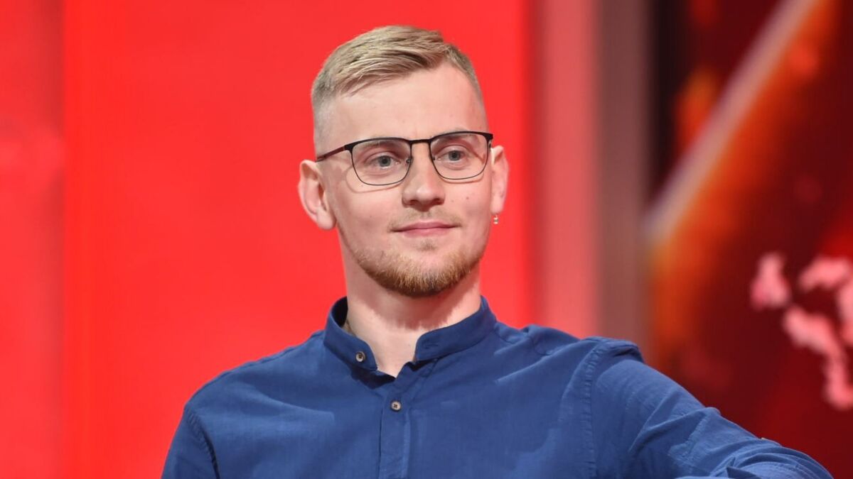 Karol, uczestnik programu Rolnik szuka żony, siedzi na tle czerwonego studia. Ma na sobie granatową koszulę i okulary, a jego fryzura jest krótka i starannie ułożona. Patrzy spokojnie przed siebie z lekkim uśmiechem.